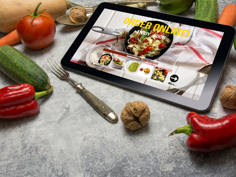 Tablet with online food shopping open and surrounded by peppers, tomatoes, and cucumbers.